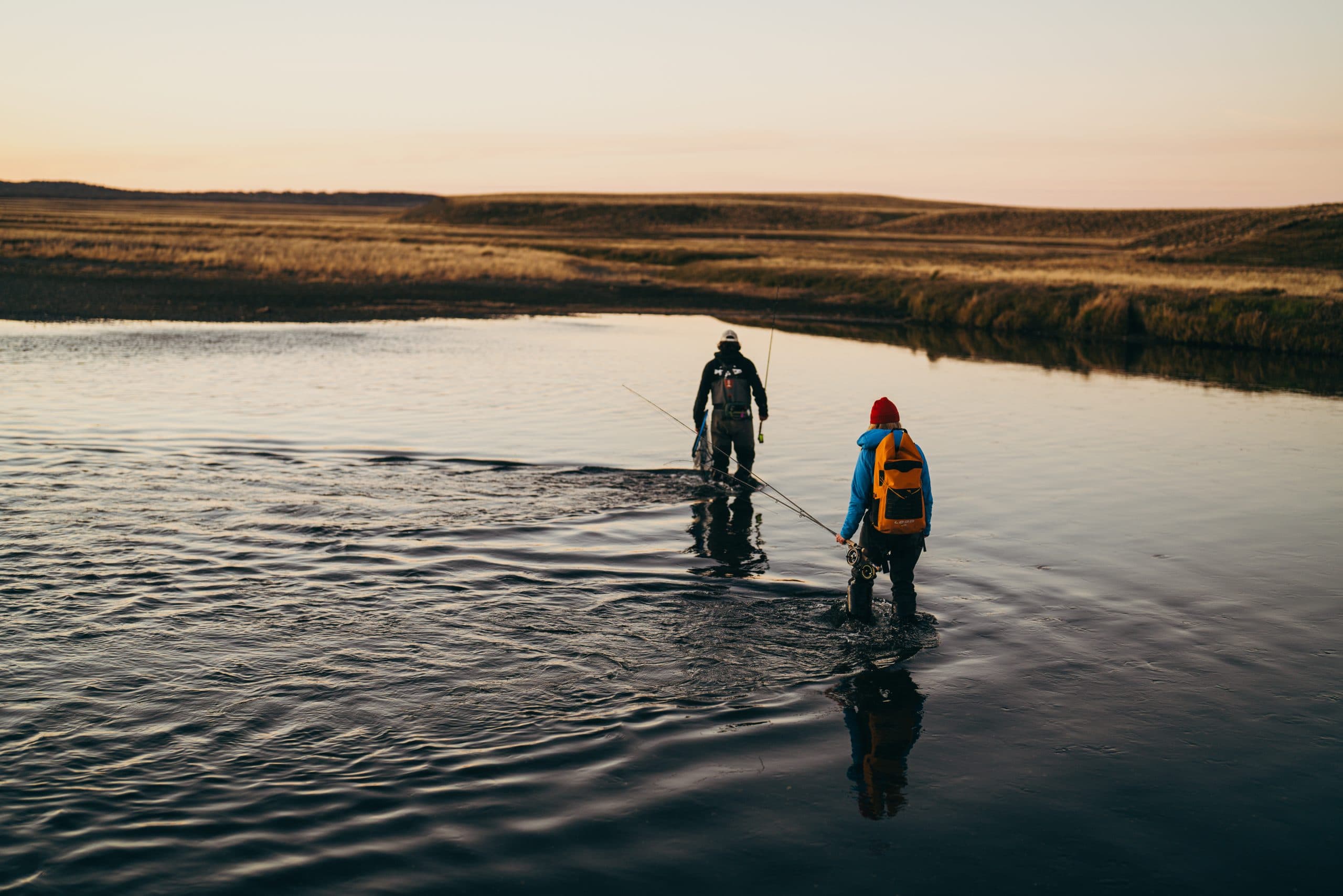 Two anglers wading across a shallow river, one is wearing a yellow Loop 25L dry backpack