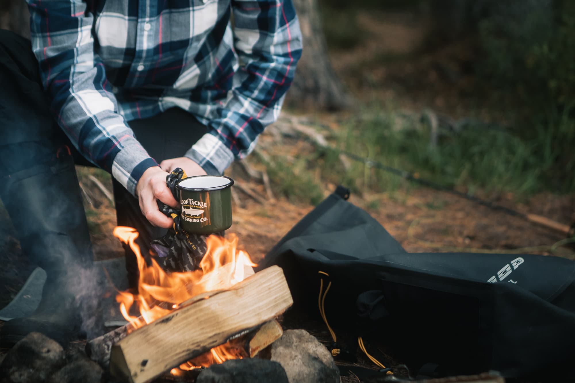 A mug of coffee being held over a campfire, the man is wearing a midnight sun shirt
